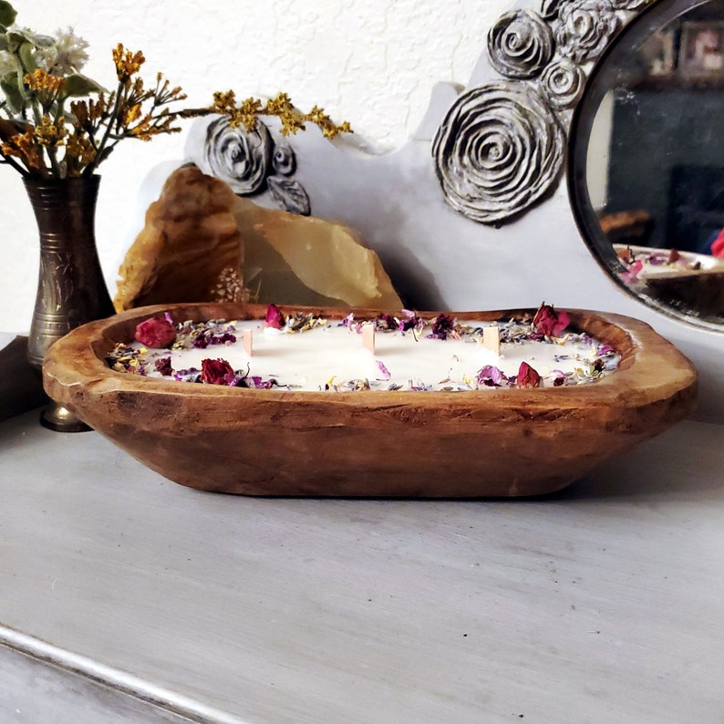 May include: A large, wooden dough bowl candle filled with white wax and three wooden wicks. The candle is decorated with dried flowers and rose petals. The bowl is brown and sits on a gray surface.