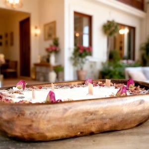 May include: A wooden candle holder filled with white wax, rose petals, and pink rose buds. The candle has several wooden wicks. The wooden bowl is oval-shaped and sits on a wooden surface. The candle is a decorative home decor item.