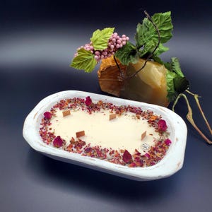 May include: A white, wooden dough bowl candle with a creamy wax interior. The candle is decorated with dried rose petals and buds, and has four wooden wicks. A decorative stone and floral arrangement are in the background.