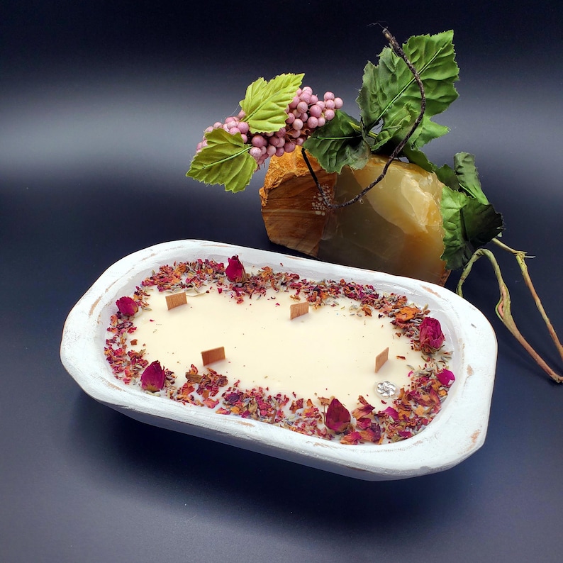 May include: A white, wooden dough bowl candle with a creamy wax interior. The candle is decorated with dried rose petals and buds, and has four wooden wicks. A decorative stone and floral arrangement are in the background.