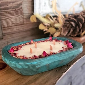 May include: A teal-colored, wooden dough bowl candle with a creamy wax interior. The candle is decorated with dried rose petals and pink rose buds. It has six wooden wicks. The candle is a decorative home fragrance item.