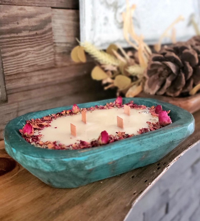 May include: A teal-colored, wooden dough bowl candle with a creamy wax interior. The candle is decorated with dried rose petals and pink rose buds. It has six wooden wicks. The candle is a decorative home fragrance item.