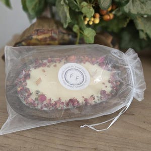 May include: A handmade candle in a wooden oval bowl, surrounded by dried rose petals. The candle is white with two wooden wicks and a circular sticker that reads "Funk Free Natural Candles". The candle is packaged in a sheer white bag.