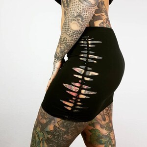 Puede incluir: Una minifalda negra con un panel lateral de recortes. La falda se muestra en una persona con tatuajes en las piernas y los brazos.