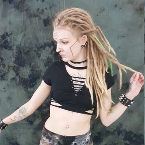 Op de afbeelding: Een persoon met lange blonde dreadlocks draagt een zwart gecropped topje met een vetersluiting en een zwart-witte tie-dye legging. Ze draagt ​​meerdere armbanden om haar polsen en heeft een neusring en oorbellen.
