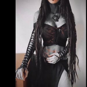 May include: A woman with long black dreadlocks is wearing a black and brown top with a lace-up detail. She is wearing black lace gloves and a silver necklace with a pendant.