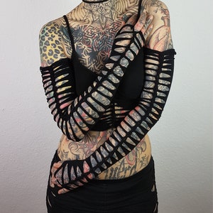 May include: A person with extensive tattoos and piercings is wearing a black crop top and a black skirt with cutouts. They are also wearing black fingerless gloves with cutouts.