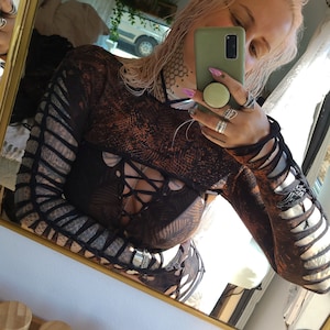 Puede incluir: Una persona lleva una camiseta de malla negra y naranja con un cierre de encaje en la parte delantera. La persona se está tomando una selfie en un espejo, sosteniendo una funda de teléfono verde con un soporte pop.