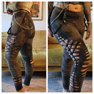 Könnte beinhalten: Dunkelgraue Leggings mit einem Distressed-Tie-Dye-Effekt. Die Leggings haben seitliche Ausschnitte. Ein schwarzes Ledergeschirr mit Kettenbesatz ist an den Leggings befestigt.