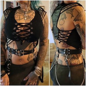 Könnte beinhalten: Ein schwarzes Crop-Top mit Schnürung vorne und einem Lederimitat-Rückenpanel. Das Top wird mit einem schwarzen Ledergürtel mit silbernen Schnallen und Ketten getragen.