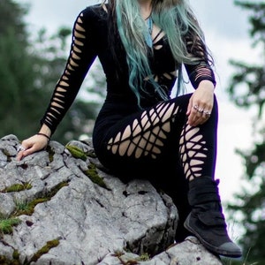 Peut inclure: Une femme aux longs cheveux argentés et aux mèches bleues porte une robe noire à manches longues et à découpes et des bottes noires. Elle est assise sur un gros rocher dans un décor de forêt.