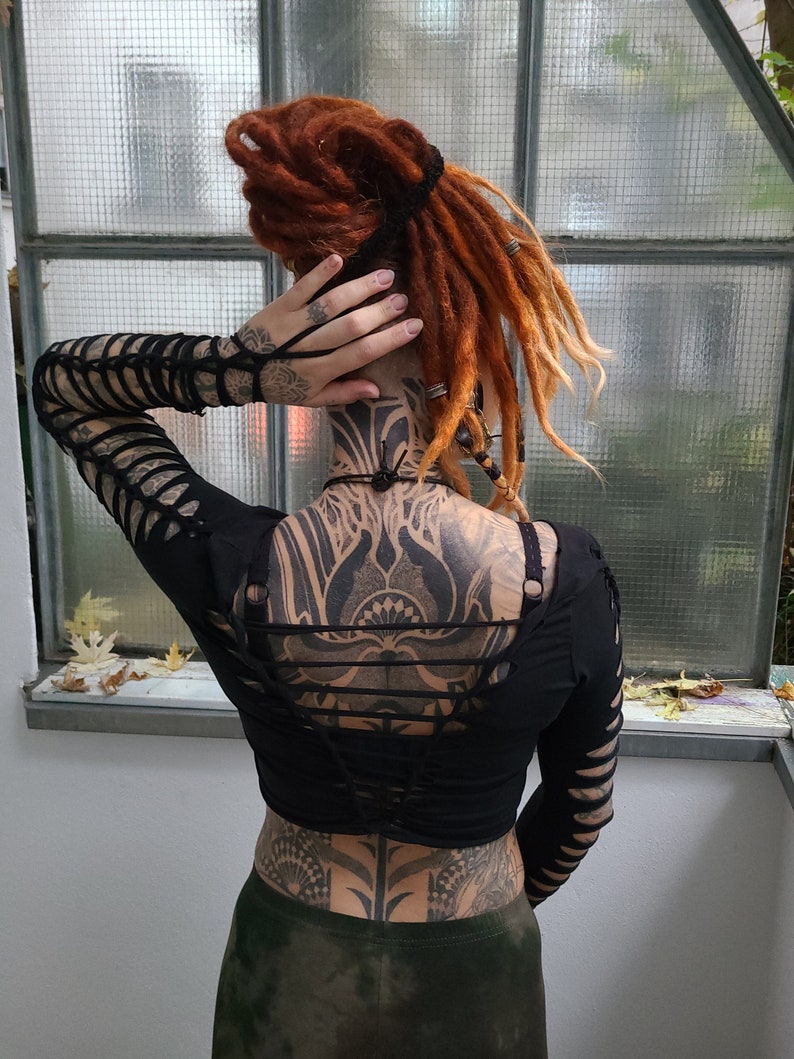 Pu&ograve; includere: Un top nero a maniche lunghe e corto con un dettaglio di pizzo sul retro. Il top &egrave; indossato sopra un design di tatuaggio a corpo intero.
