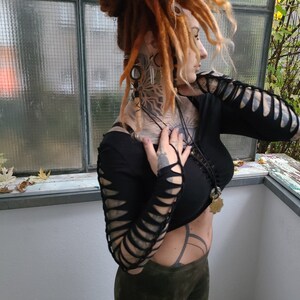 Pu&ograve; includere: Una donna con dreadlock rossi indossa una camicia nera a maniche lunghe con dettagli ritagliati sulle maniche. Indossa pantaloni verdi e ha diversi collane e orecchini.