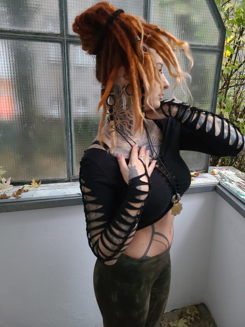 Pu&ograve; includere: Una donna con dreadlock rossi indossa una camicia nera a maniche lunghe con dettagli ritagliati sulle maniche. Indossa pantaloni verdi e ha diversi collane e orecchini.