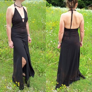 May include: A black halter maxi dress with a deep V-neck and a high slit on the left side. The dress has a flowing silhouette and a halter tie. The dress is worn outdoors in a field of green grass and wildflowers.