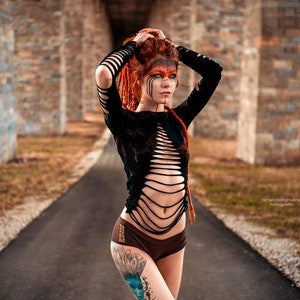 Op de afbeelding: Een vrouw met rood haar en oranje en blauwe gezichtsverf draagt een zwart, gescheurd, lang mouw shirt en een bruine broek. Ze heeft een tatoeage op haar been.