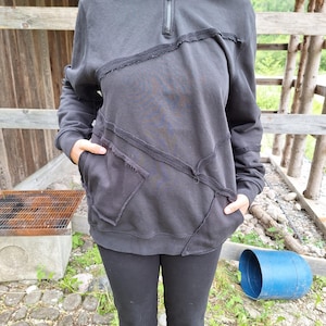 Może przedstawiać: Czarna bluza z kapturem z zapięciem na pół zamek i unikalnym wzorem patchworku. Bluza posiada kaptur, długie rękawy i przednią kieszeń. Projekt zawiera widoczne szwy i surowe krawędzie, nadając jej postarzany wygląd. Osoba nosi również czarne spodnie.