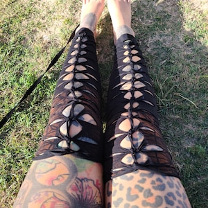 May include: A pair of black leggings with lace-up details on the sides. The leggings are worn by a person with tattoos on their legs, including a floral design and a leopard print.