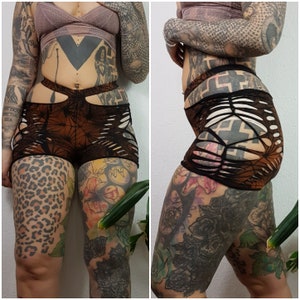 Könnte beinhalten: Braune und schwarze Shorts mit einem Distressed-Design. Die Shorts haben Ausschnittdetails und eine hohe Taille. Der Stoff hat ein Muster. Die Shorts werden von vorne und von der Seite gezeigt.