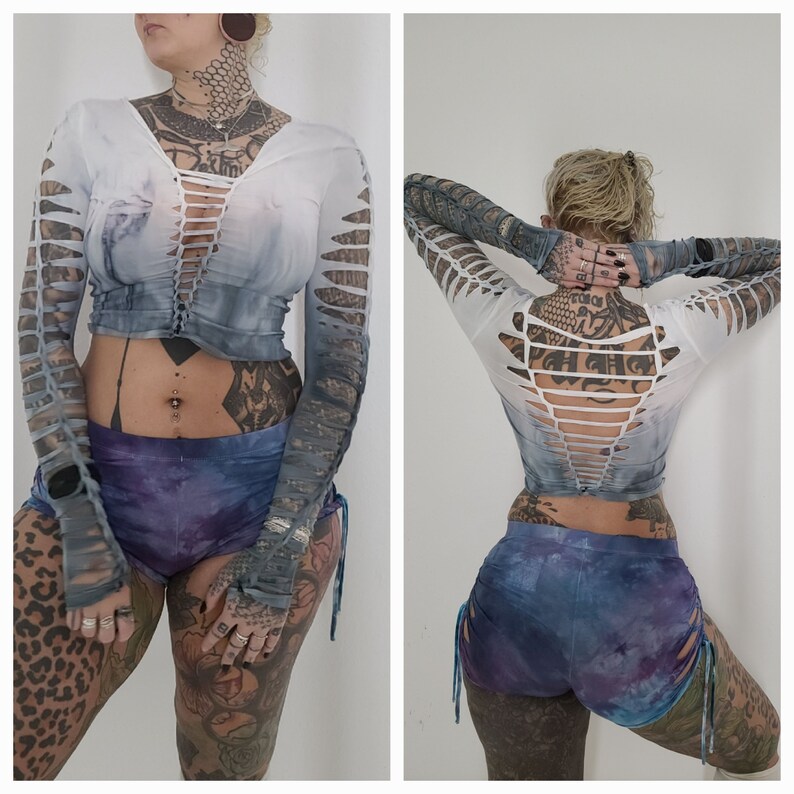 Pu&ograve; includere: Un top bianco a maniche lunghe con effetto tie-dye e dettagli tagliati. Il top ha una chiusura con lacci sul davanti e uno scollo a V sul retro. La modella indossa il top con un paio di pantaloncini tie-dye blu e viola.