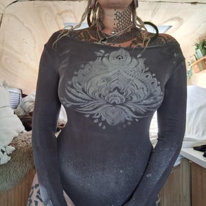 Peut inclure: Un body à manches longues gris foncé avec un grand motif floral gris clair sur le devant. Le body a une encolure dégagée et des manches évasées. Le tissu a un aspect vieilli et délavé.