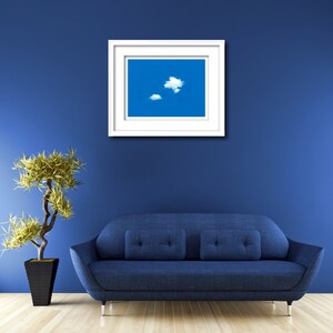 May include: A white picture frame with a blue sky and white clouds image inside. The frame is hanging on a blue wall above a blue sofa.