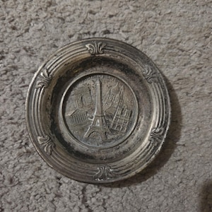 May include: A round silver-toned metal decorative dish featuring an embossed relief of Paris landmarks including the Eiffel Tower, the Arc de Triomphe, and Notre-Dame Cathedral. The rim has a raised border with decorative floral-like accents.