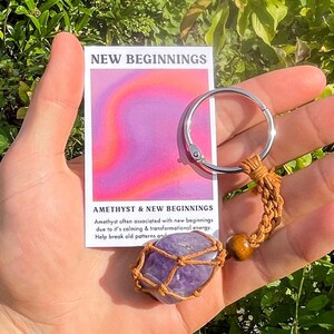May include: Amethyst crystal keychain with a brown leather cord and a wooden bead. The keychain is wrapped in a brown macrame net. The card reads "New Beginnings" and "Amethyst & New Beginnings". Amethyst often associated with new beginnings due to it's calming & transformational energy. Help break old patterns and