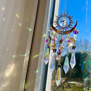 May include: A celestial-themed suncatcher with a brass-colored sun and crescent moon design. It features hanging crystals, stars, and iridescent beads. The suncatcher is suspended by a delicate chain, creating a decorative and eye-catching piece.