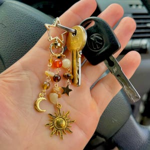 Może przedstawiać: Złoty brelok do kluczy z zawieszką w kształcie gwiazdy, księżyca i słońca, a także różnymi koralikami w odcieniach brązu, pomarańczy i kremu. Brelok jest przymocowany do dwóch kluczyków do samochodu. Tło przedstawia kierownicę samochodu.