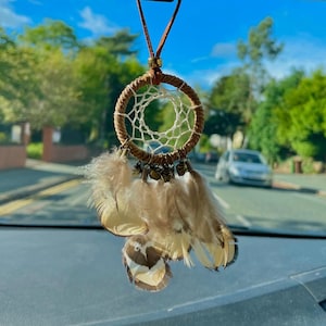 Puede incluir: Un atrapasueños marrón con red blanca y plumas beige cuelga del espejo retrovisor de un coche. El atrapasueños tiene pequeñas campanas adjuntas.