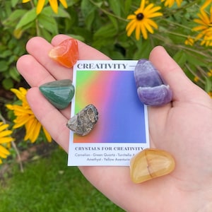 May include: A set of five crystals for creativity, including carnelian, green quartz, turquoise, amethyst, and yellow aventurine. The crystals are arranged on a hand with a card that reads "CREATIVITY - CRYSTALS FOR CREATIVITY".