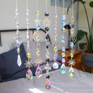 Pode incluir: Um conjunto de sete apanhadores de sol de cristal com temas celestiales e religiosos. Cada apanhador de sol apresenta um design diferente, incluindo uma lua crescente, uma estrela, um sol, uma cruz e um pentagrama. Os apanhadores de sol são feitos de cristal transparente e têm cores arco-íris iridescentes.