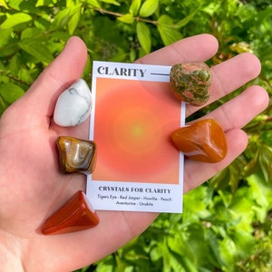 May include: A hand displaying a selection of crystals and a card. The card is labelled "Clarity" with an orange and yellow gradient. The crystals include Tiger's Eye, Red Jasper, Howlite, Peach Aventurine, and Unakite.