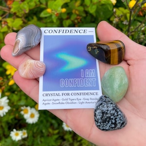 May include: A set of crystals for confidence, including apricot agate, gold tiger's eye, grey banded agate, snowflake obsidian, and light aventurine. The crystals are arranged on a hand with a card that reads "CONFIDENCE - I AM CONFIDENT. CRYSTAL FOR CONFIDENCE."