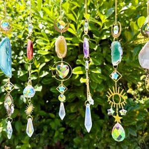 May include: A collection of hanging crystal suncatchers with gold-colored chains and accents. Each suncatcher features a unique combination of crystals, gemstones, and decorative charms, including teardrops, stars, and sunbursts. The background is a lush green bush.