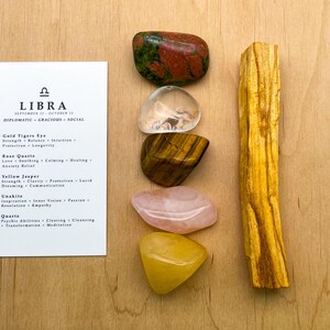 May include: A set of crystals for Libra zodiac sign, including gold tiger's eye, rose quartz, yellow jasper, unakite, clear quartz, and a palo santo stick. The crystals are arranged in a vertical stack on a wooden surface. The card lists the crystals and their associated properties.