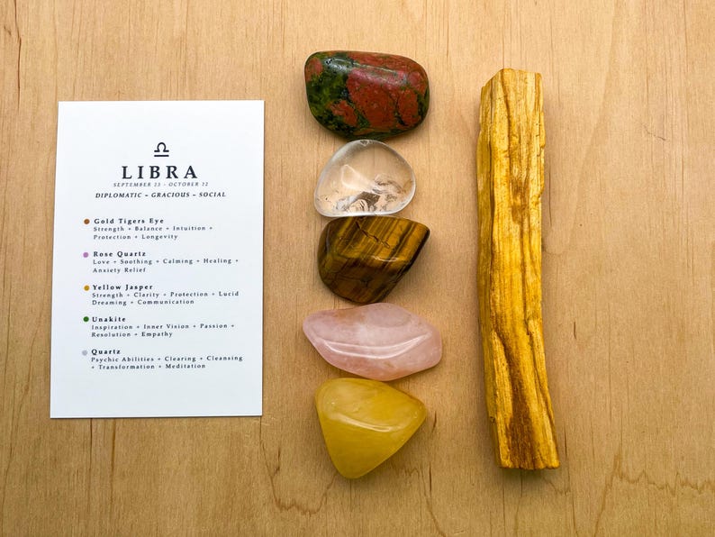May include: A set of crystals for Libra zodiac sign, including gold tiger's eye, rose quartz, yellow jasper, unakite, clear quartz, and a palo santo stick. The crystals are arranged in a vertical stack on a wooden surface. The card lists the crystals and their associated properties.