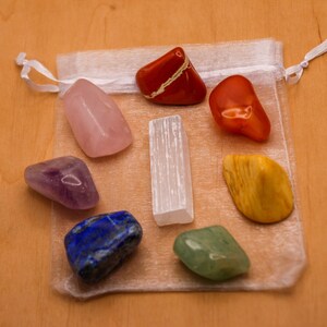 May include: A set of seven chakra stones in a white organza bag. The stones are arranged in a circle and include amethyst, rose quartz, clear quartz, citrine, green aventurine, lapis lazuli, and red jasper.