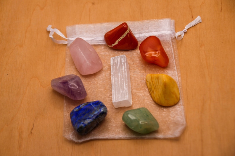 May include: A set of seven chakra stones in a white organza bag. The stones are arranged in a circle and include amethyst, rose quartz, clear quartz, citrine, green aventurine, lapis lazuli, and red jasper.