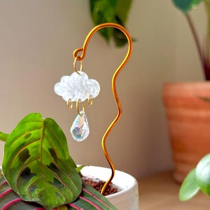 May include: A decorative plant stake featuring a wavy gold-toned metal wire topped with a white, textured cloud charm. Three small gold rings hang from the cloud, with a single clear, faceted, iridescent teardrop crystal suspended from the center.
