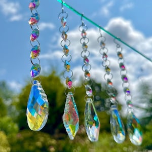 May include: Several strands of iridescent crystal suncatchers hang against a backdrop of a blue sky and green foliage. Each strand features a series of small, round crystals connected by silver-colored rings, culminating in a larger, teardrop-shaped crystal.