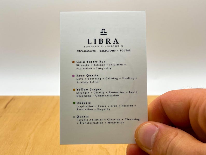 May include: A white card with the astrological sign Libra and the text "SEPTEMBER 23 - OCTOBER 22" and "DIPLOMATIC - GRACIOUS - SOCIAL". The card lists five crystals associated with Libra: Gold Tiger's Eye, Rose Quartz, Yellow Jasper, Unakite, and Quartz. Each crystal is listed with its properties.