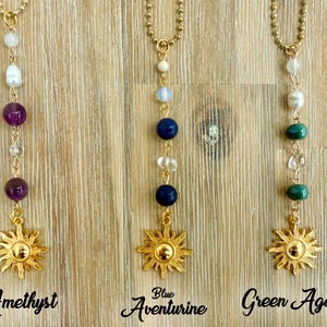 Puede incluir: Tres collares dorados con diferentes combinaciones de piedras preciosas y colgantes de sol. Los collares est&aacute;n etiquetados como "Amatista", "Aventurina azul" y "&Aacute;gata verde". Cada collar tiene una cadena dorada y un colgante en forma de sol.