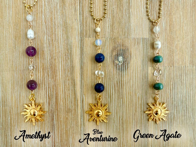 Puede incluir: Tres collares dorados con diferentes combinaciones de piedras preciosas y colgantes de sol. Los collares est&aacute;n etiquetados como "Amatista", "Aventurina azul" y "&Aacute;gata verde". Cada collar tiene una cadena dorada y un colgante en forma de sol.