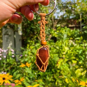 May include: A red gemstone wrapped in brown macrame cord with a wooden bead and a silver key ring. The keychain is held in a hand against a blurred background of green foliage.