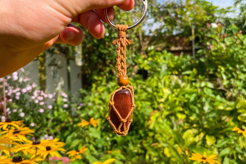 May include: A red gemstone wrapped in brown macrame cord with a wooden bead and a silver key ring. The keychain is held in a hand against a blurred background of green foliage.