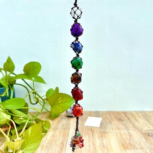 May include: A hanging chakra stone ornament featuring seven colorful gemstones, each encased in a knotted brown cord. The stones are purple, blue, green, brown, red, and orange. The ornament is suspended from a black cord and has a cluster of small stones at the bottom.