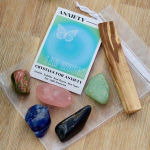 May include: A set of crystals for anxiety, including unakite, sodalite, rose quartz, blue tiger's eye, and light aventurine, along with a palo santo stick and a small white pouch. The card reads "Anxiety I Am Enough Crystals for Anxiety".