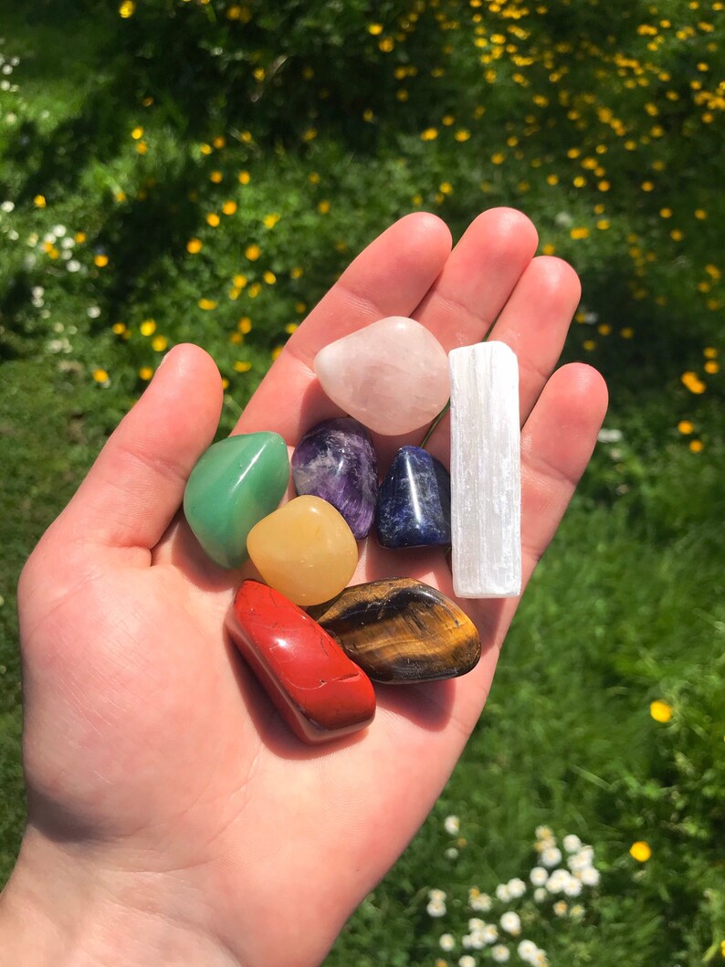 May include: A hand holding seven crystals: a green aventurine, a yellow citrine, a red carnelian, a tiger's eye, a blue sodalite, a purple amethyst, and a white selenite stick.